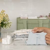 Person folding a shirt on a table with flowers and laundry supplies in the background