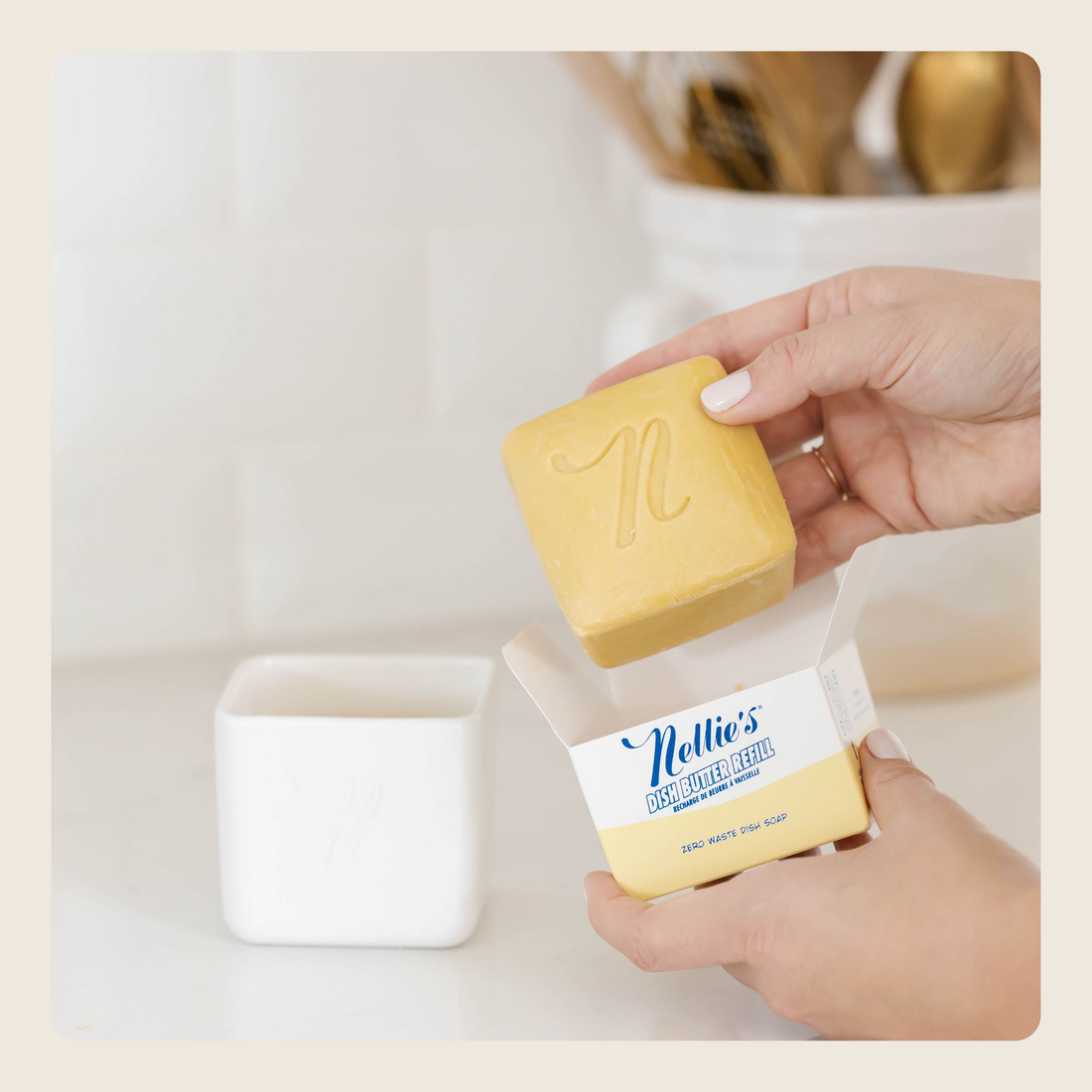 A person holds a yellow soap bar above its white and yellow Nellie's Soap dish soap box, with a white soap dish sitting on a white countertop in a bright kitchen setting.