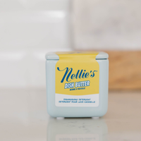 A light blue plastic container of Nellie's Dish Butter, featuring a yellow label with blue script text, set against a softly blurred kitchen background.