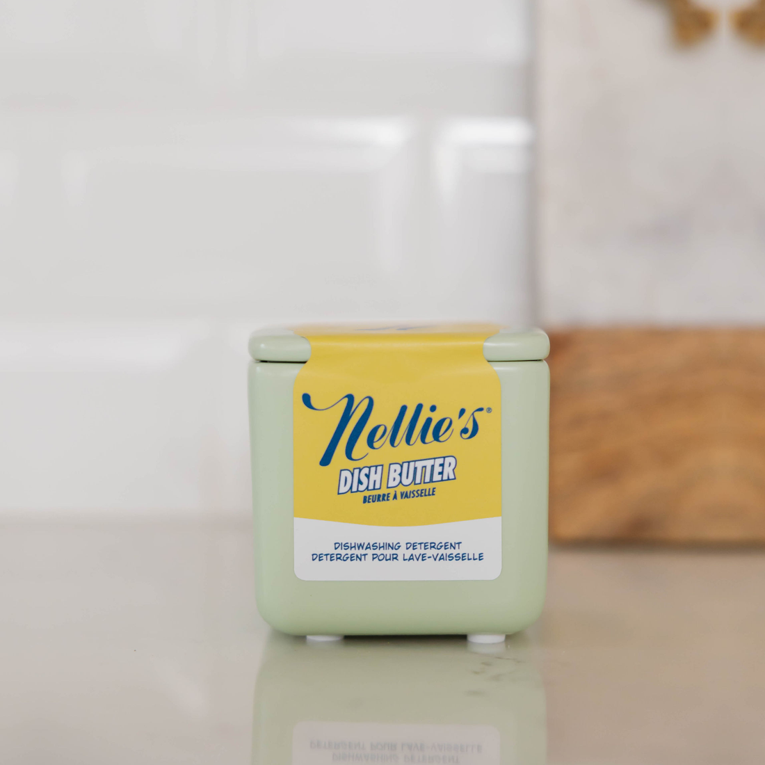 A small square container of Nellie's Dish Butter dishwashing detergent with a yellow label and pale green container, sitting on a reflective light surface with a blurred kitchen background.