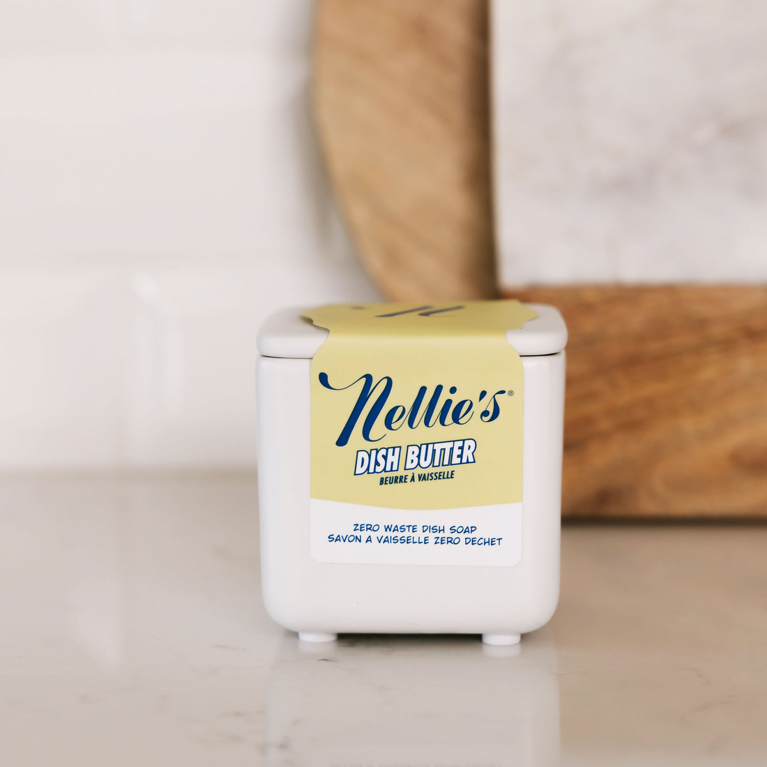 A white plastic container of Nellie's Dish Butter with a pale yellow label, set on a light marble countertop with a blurred wooden cutting board and white subway tiles in the background.