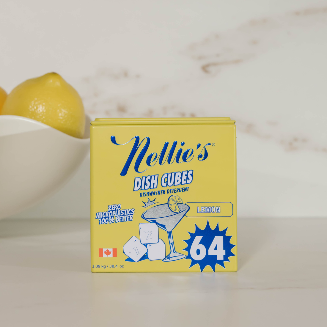 A yellow box of Nellie's Dish Cubes Lemon flavor dishwasher detergent stands on a light countertop, with a white bowl and part of a lemon blurred in the background.