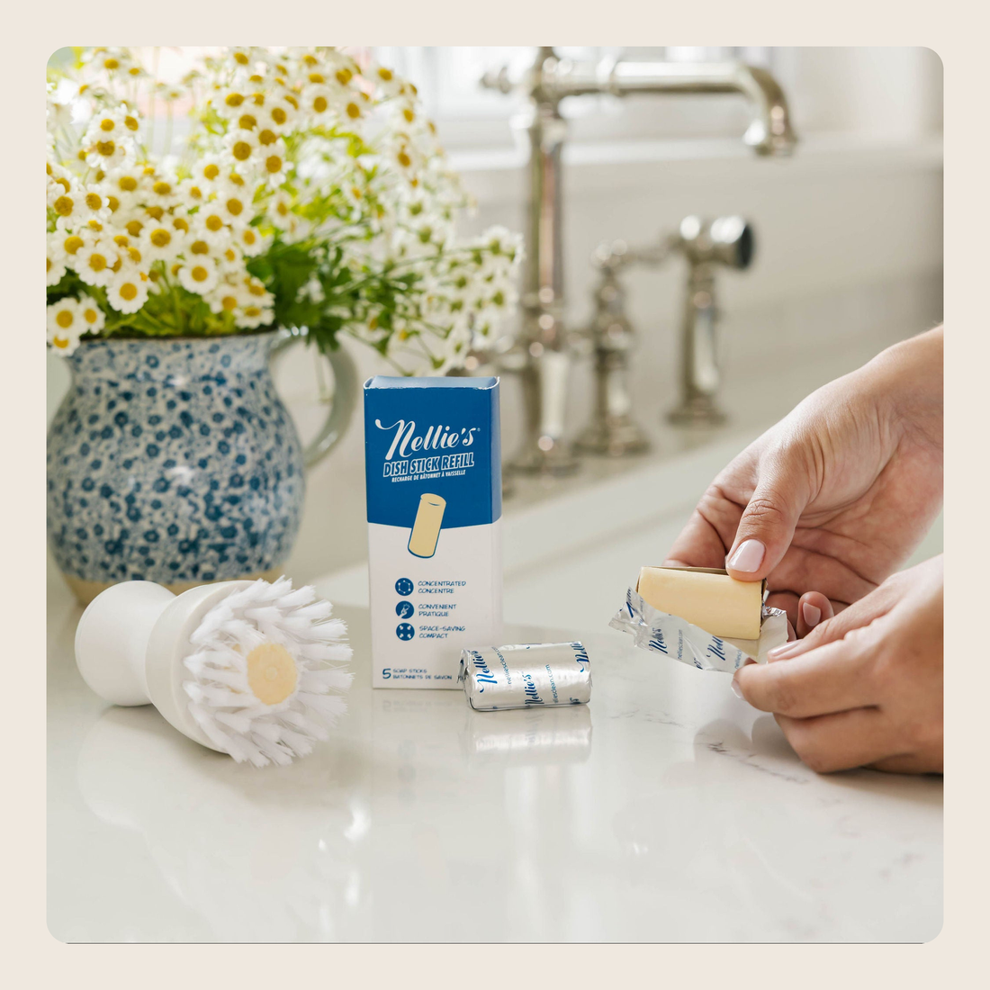 A person's hands are shown applying Nollie's Pain Relieving Band-Aid cream to a bandage. The product box, a white cleaning brush, and a silver foil-wrapped bandage are arranged on a white kitchen counter in front of a blurred sink and a vase of white daisies.