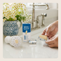 A person's hands are shown applying Nollie's Pain Relieving Band-Aid cream to a bandage. The product box, a white cleaning brush, and a silver foil-wrapped bandage are arranged on a white kitchen counter in front of a blurred sink and a vase of white daisies.