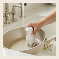 A hand uses a white and silver cleaning brush with soft bristles to scrub the inside of a beige frying pan under running water in a sink.