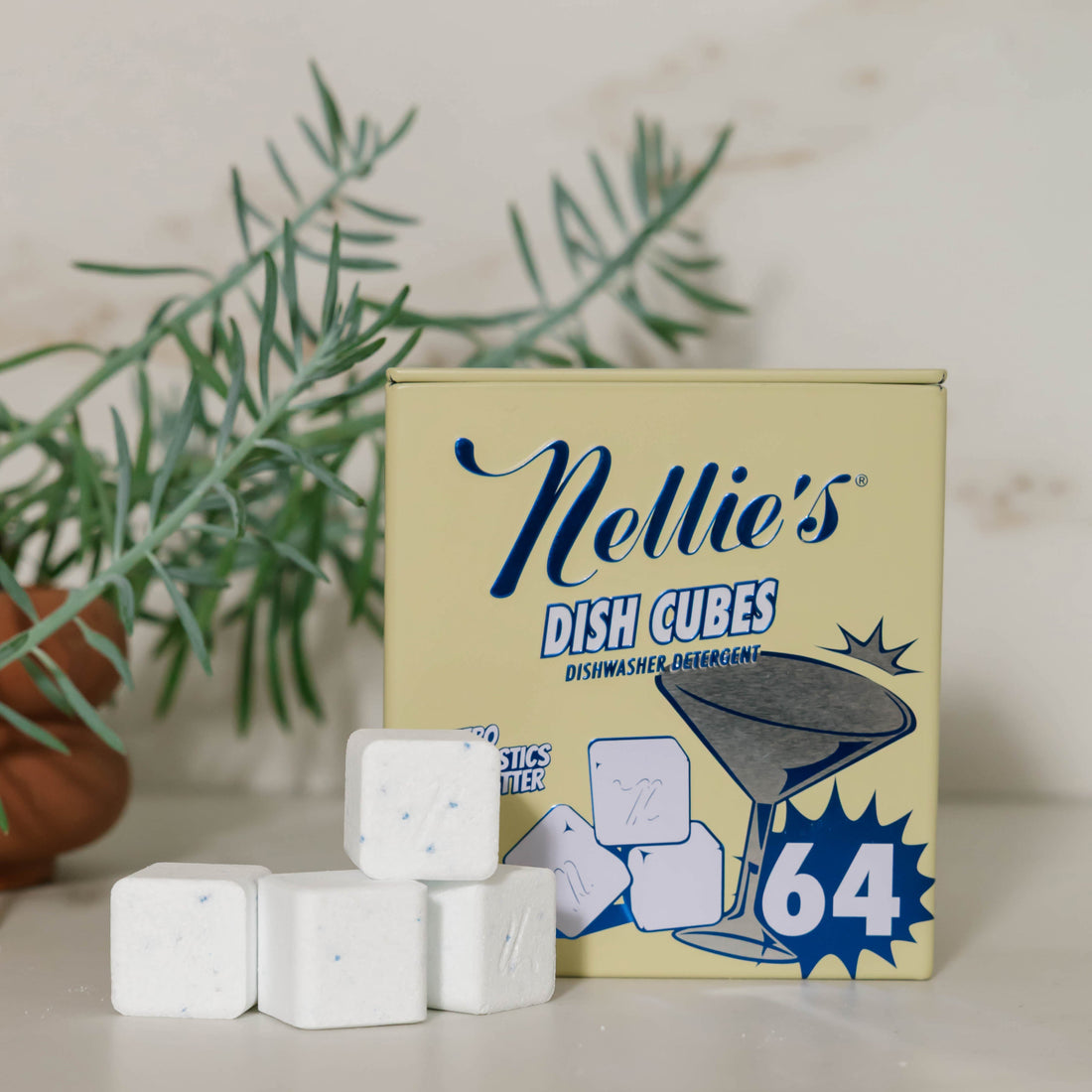 A box of Nellie's Dish Cubes with several white cube-shaped cleaning blocks placed in front, styled on a light surface with a blurred green plant in the background.