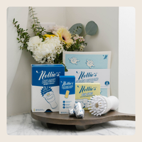 Nellie's dishwashing products displayed on a wooden tray, including a blue and white training pouch, a small packet, foil-wrapped items, a white silicone training brush, and a booklet, styled with flowers in the background.