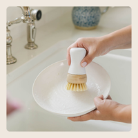 A close-up of a hand holding a white and natural wood dishwashing brush against a white ceramic bowl in a sink, with a silver faucet visible in the background.