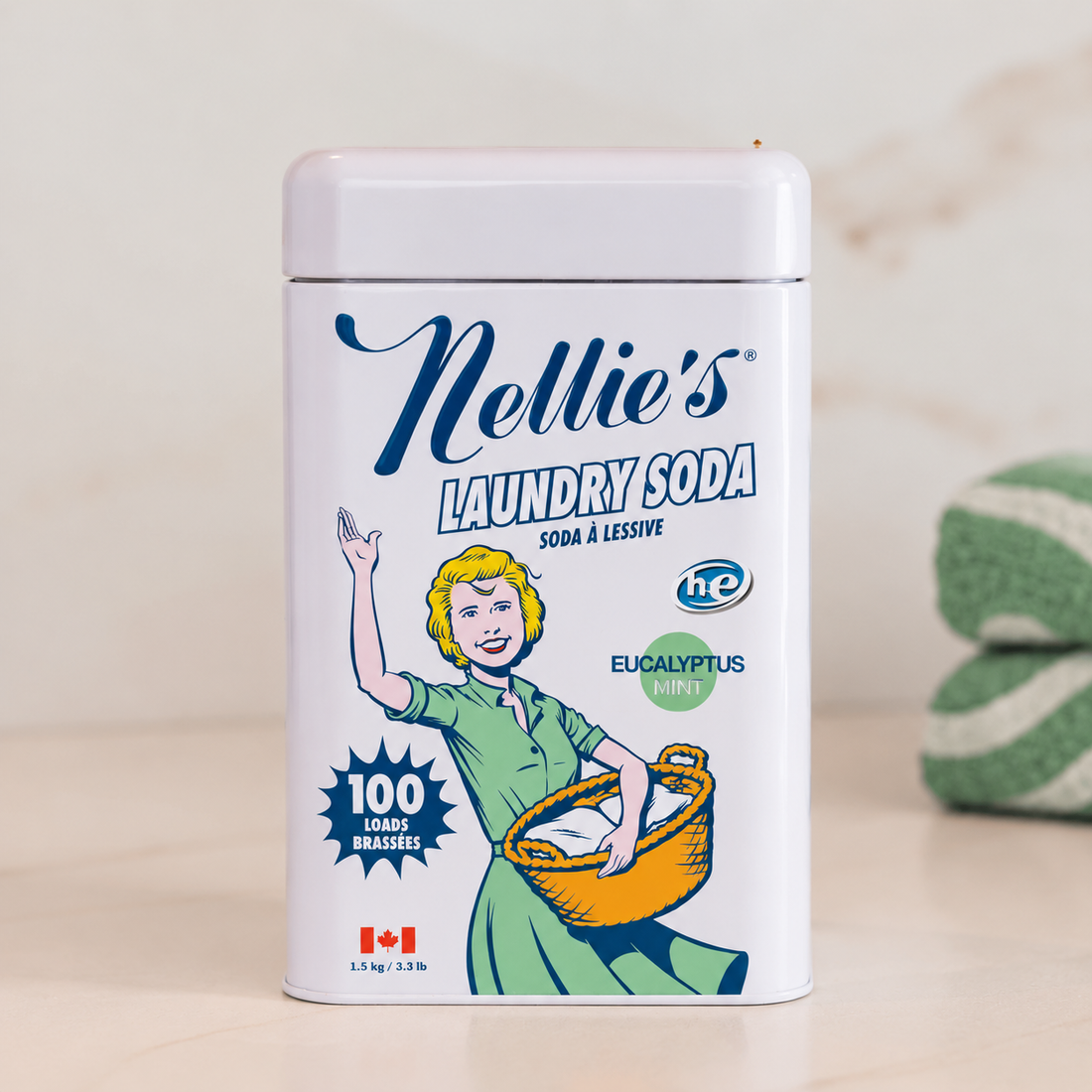 A white rectangular tin of Nellie's Laundry Soda, SODA à Lessive, with a vintage-style illustration of a woman in a green dress holding a basket. The label includes '100 Laves Brésiles' and 'Eucalyptus Mint' in blue and green text.