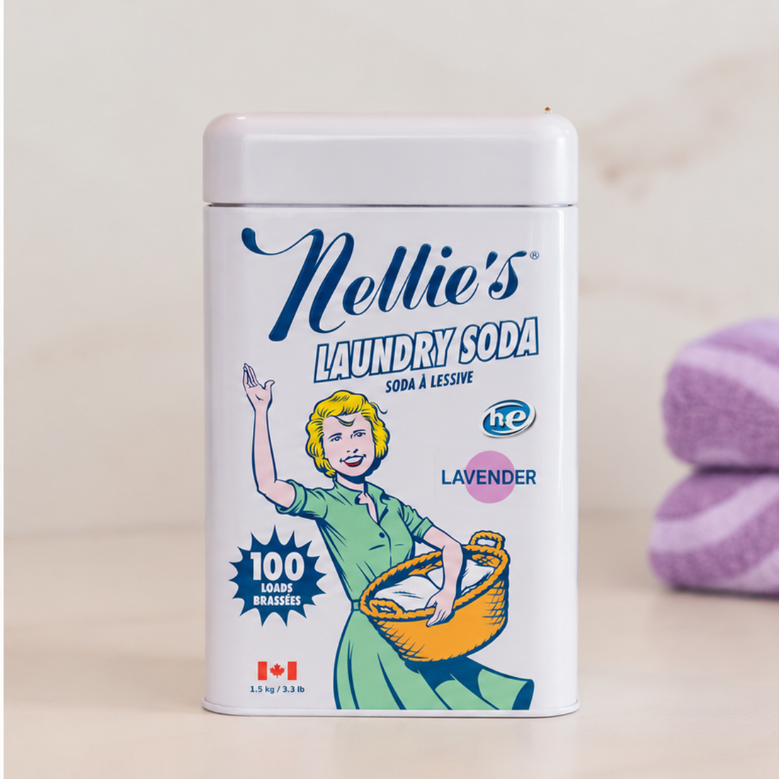A white plastic tin of Nellie's Laundry Soda in Lavender scent, featuring a vintage-style illustration of a woman holding a laundry basket. The tub is set on a light beige surface with a soft-focus background showing a purple towel.