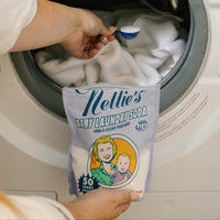 A person adds Nellie's Baby Laundry Soda to a washing machine drum using a blue measuring scoop. The product packaging is held in front of the open washing machine.