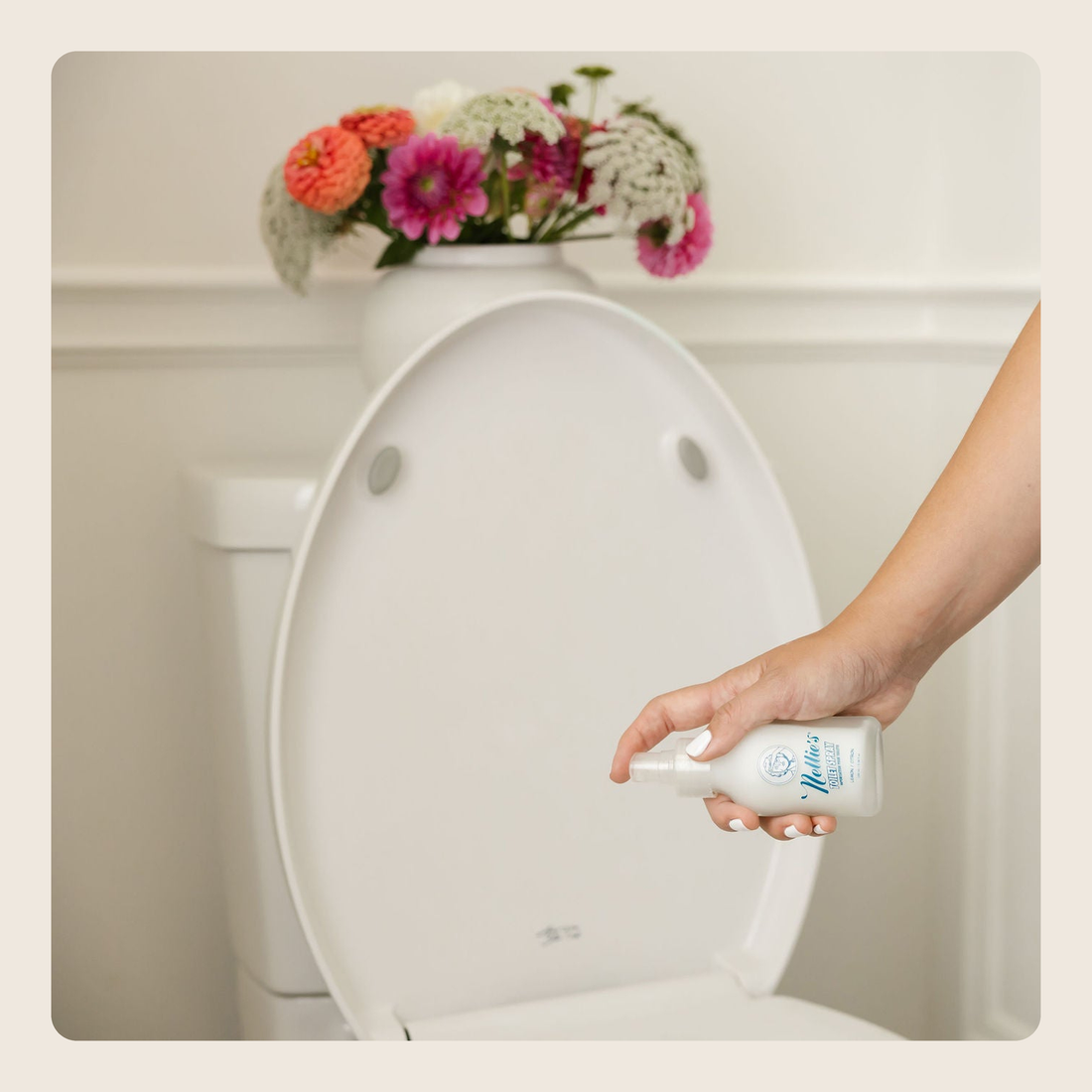 A hand holding a white spray bottle with blue text, spraying a toilet seat in a clean bathroom setting with a vase of flowers on top.