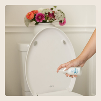 A hand holding a white spray bottle with blue text, spraying a toilet seat in a clean bathroom setting with a vase of flowers on top.